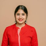 Portrait of a smiling woman wearing a red dress, earrings, and lipstick against a beige background.